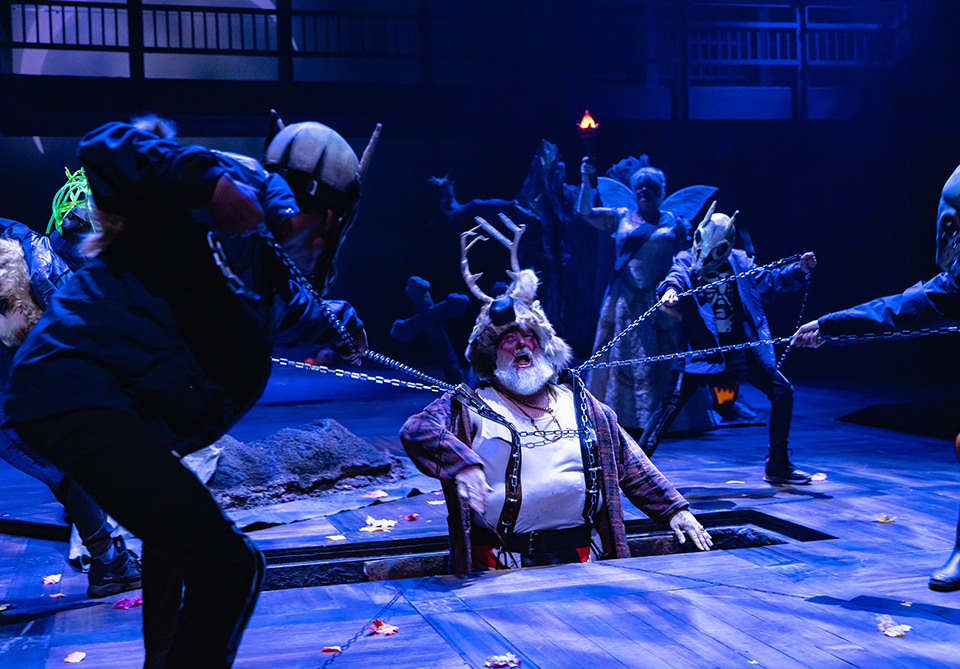 A theatrical scene with costumed performers pulling chains on a man emerging from a trapdoor, wearing antlers, on a dim stage.