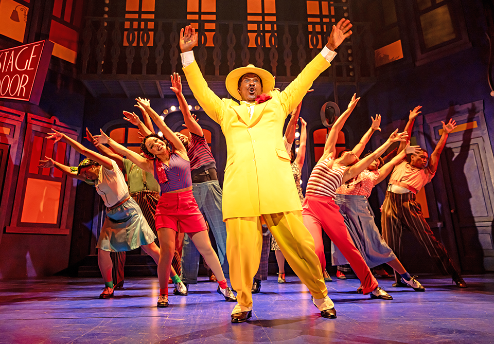 Musical theater performers dance onstage around a singer in a bright yellow suit and hat, beneath warm red and orange lighting.