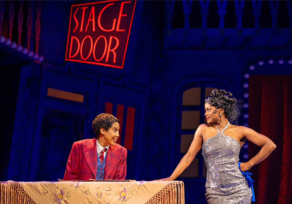 Two performers act onstage beside a floral-draped table beneath a glowing “STAGE DOOR” sign.