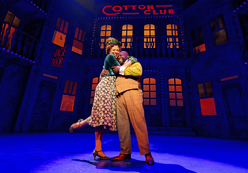 Two smiling theater performers embrace on a blue-and-orange-lit stage beneath a “COTTON CLUB” sign.