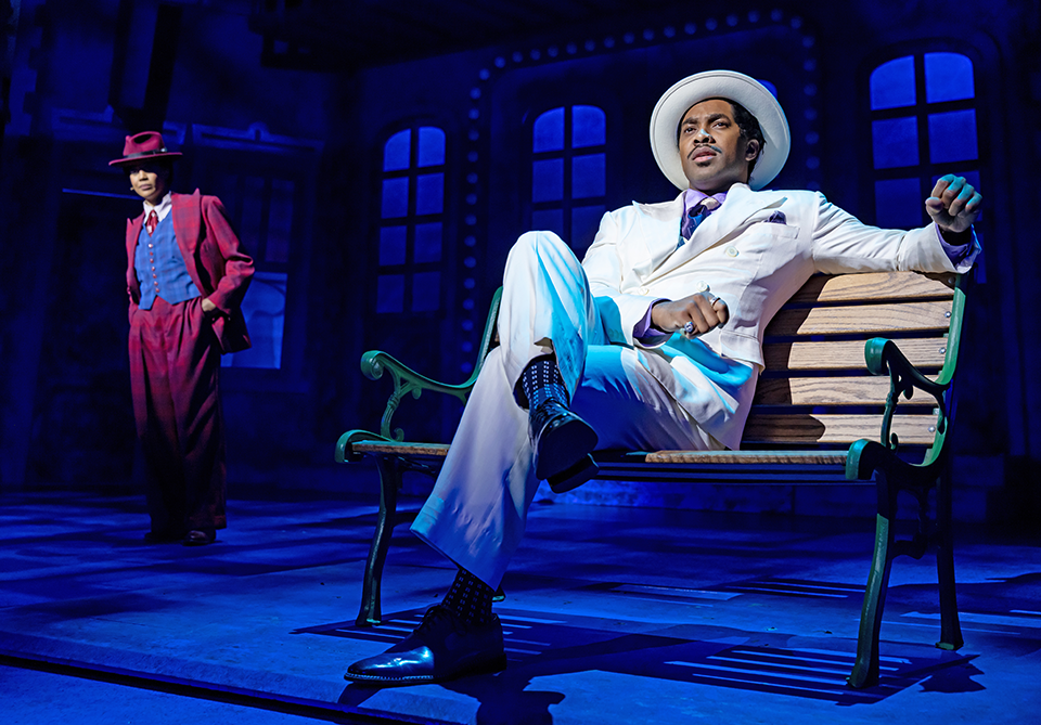 Two performers on a blue-lit stage: one in a white suit reclines on a bench while another in a burgundy suit stands behind him.