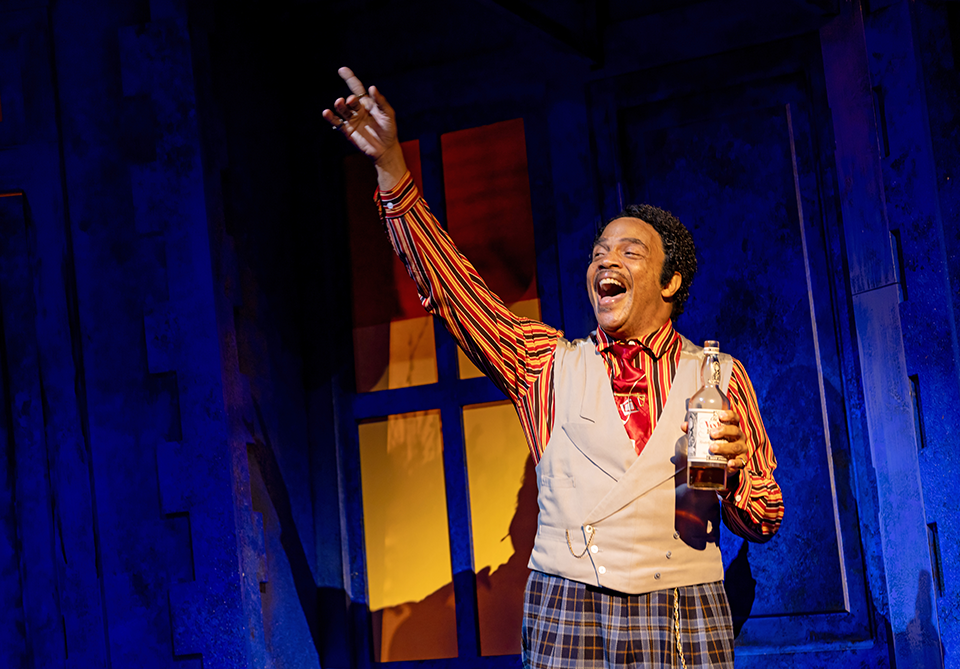Theater performer in a striped shirt and vest raises one hand while holding a bottle on a blue-lit stage.