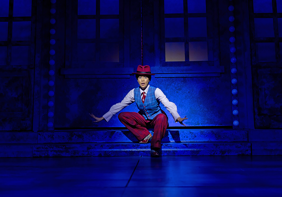 Performer in a red hat and colorful vest sits on blue-lit stage steps with arms extended.