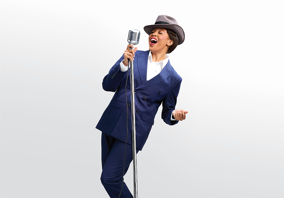 Woman in a navy suit and gray fedora singing into a vintage microphone.