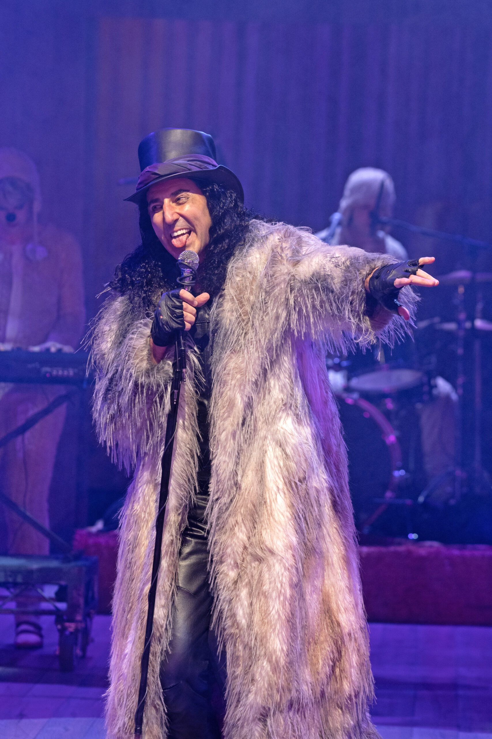  Sean Fortunato as Autolycus sings into a standing microphone while wearing a floor length fur coat and top hat